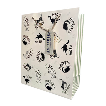 A cream gift bag with black cat illustrations and text like 'MEOW', 'STRETCH', and 'POUNCE' and golf foiling details, tied with a rope handle and a small gift tag reading 'BATTERSEA'.