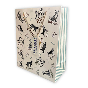 A decorative gift bag with a cream background printed with black dog illustrations, dog-related words like FETCH, WAGGING, and WOOF, rope handles, and a hanging tag reading BATTERSEA. The bag has a striped side panel in mint green and white.