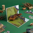 Load image into Gallery viewer, 3D Highland Cow Pop Up Card on a green backdrop, half closing, with some props surrounding it.