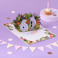 Load image into Gallery viewer, 60th birthday card for her by Cardology - floral theme for mothers, mums, sisters, aunts, friends and more - lifestyle image of the card open with a cake, bunting and confetti surrounding it
