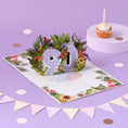 Load image into Gallery viewer, 90th birthday pop up card for her - floral theme with butterflies, birds and flowers - lifestyle image of the card open surrounded by cake, bunting and confetti
