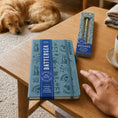 Load image into Gallery viewer, A light blue BATTERSEA notebook with a dark blue elastic band and matching blue ribbon sits on a wooden table, next to its product box. A person's hand rests near the notebook, and a golden retriever is visible in the background on a carpeted floor.
