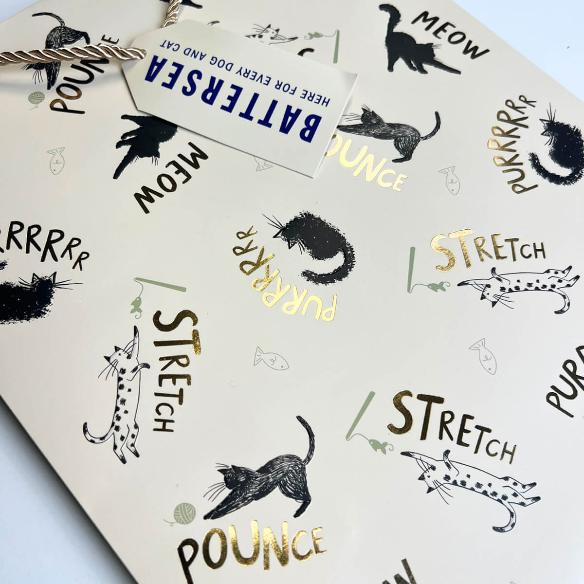 A close-up of a cat-themed gift bag black cat illustrations and gold foiled lettering with words 'MEOW', 'STRETCH', and 'POUNCe' in black and gold foil, and a white paper tag reading 'BATTERSEA HERE FOR EVERY DOG AND CAT'