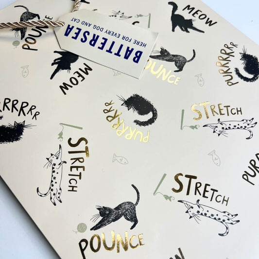 A close-up of a cat-themed gift bag black cat illustrations and gold foiled lettering with words 'MEOW', 'STRETCH', and 'POUNCe' in black and gold foil, and a white paper tag reading 'BATTERSEA HERE FOR EVERY DOG AND CAT'