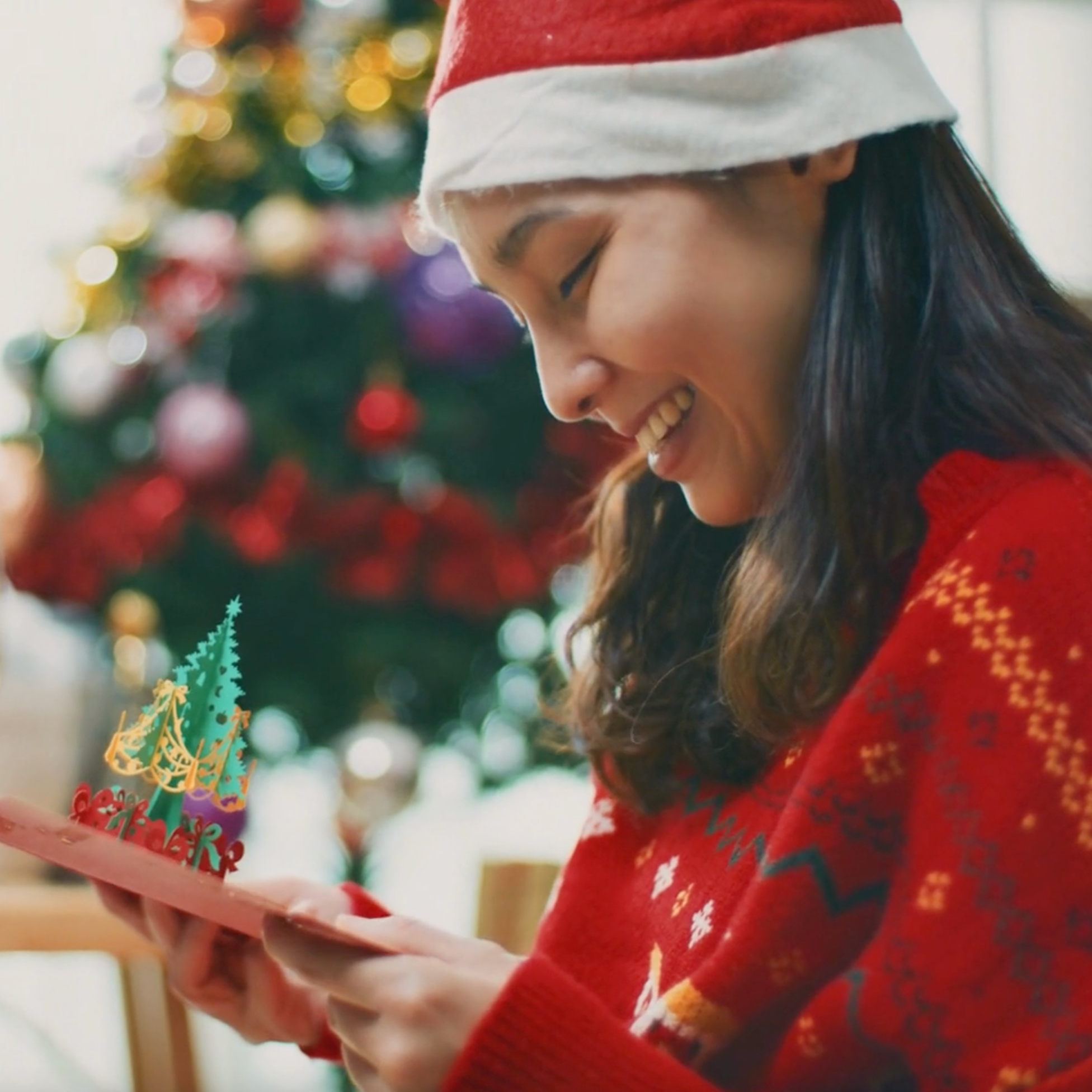 Woman in a red sweater and Santa hat holding a small Christmas tree decoration in front of a decorated Christmas tree.