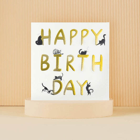 Lifestyle Image of Happy Birthday Cats Card. White with gold foiling with lots of black cats perching on the letters.