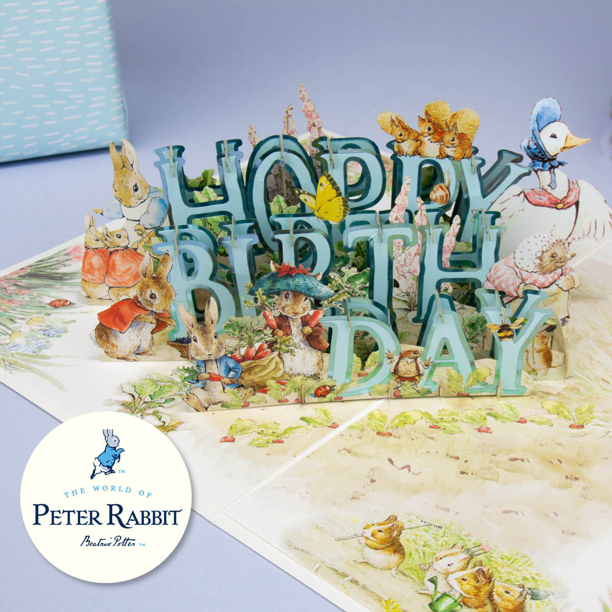 Peter Rabbit Birthday Card close up lifestyle image.  Blue 'Hoppy Birthday' surrounded by all characters from the iconic Beatrix Potter tale